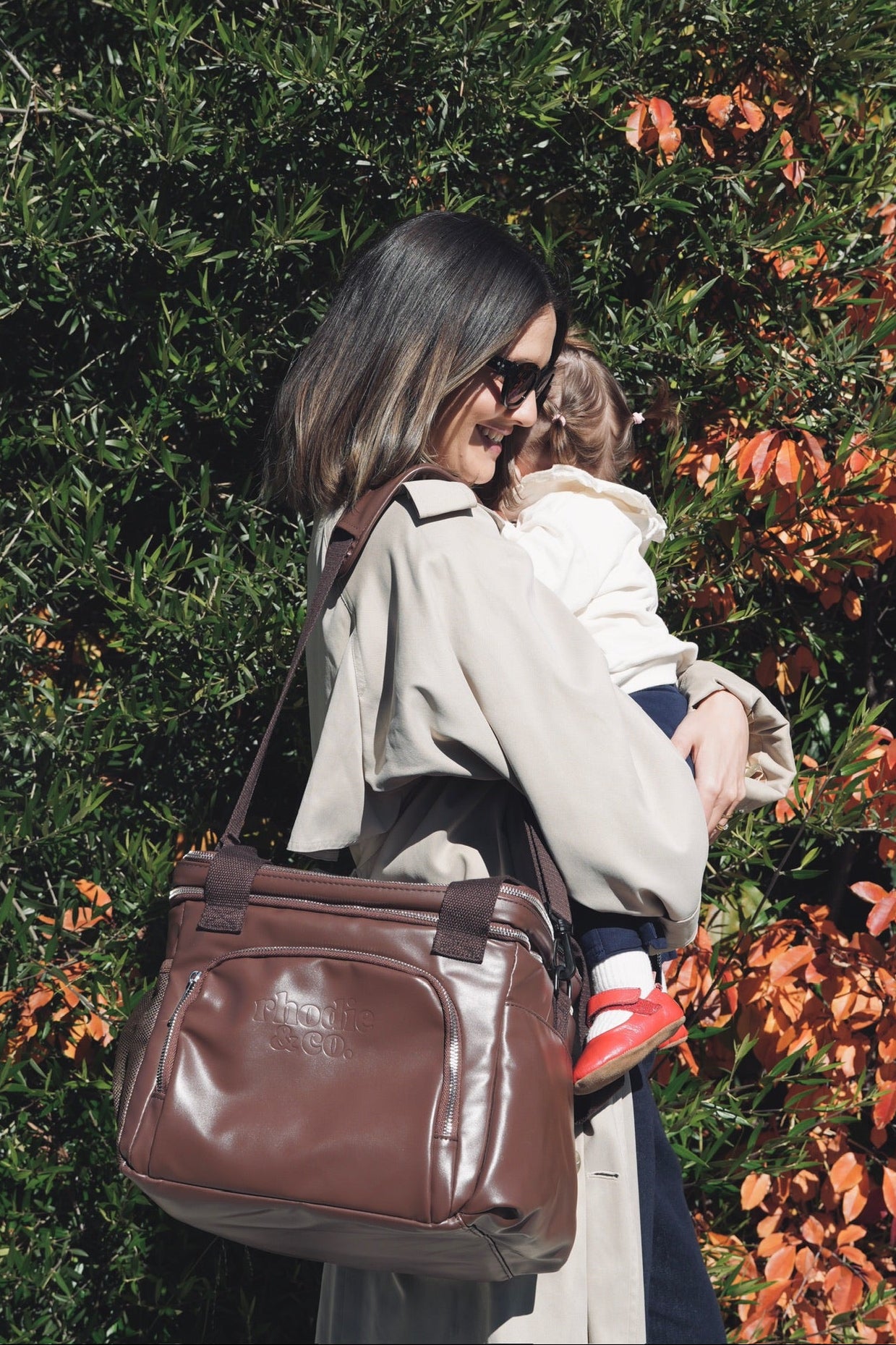 Woman holding a child and a brown thermoelectric bag in front of green and orange foliage